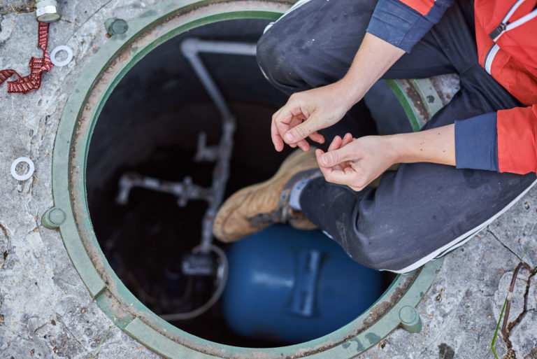 Young plumber or technician sitting on opened hatch of system of water filtration on the street in the home yard